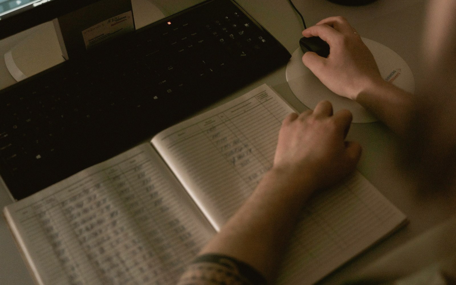 A hand rests on a ledger book beside a keyboard and computer mouse